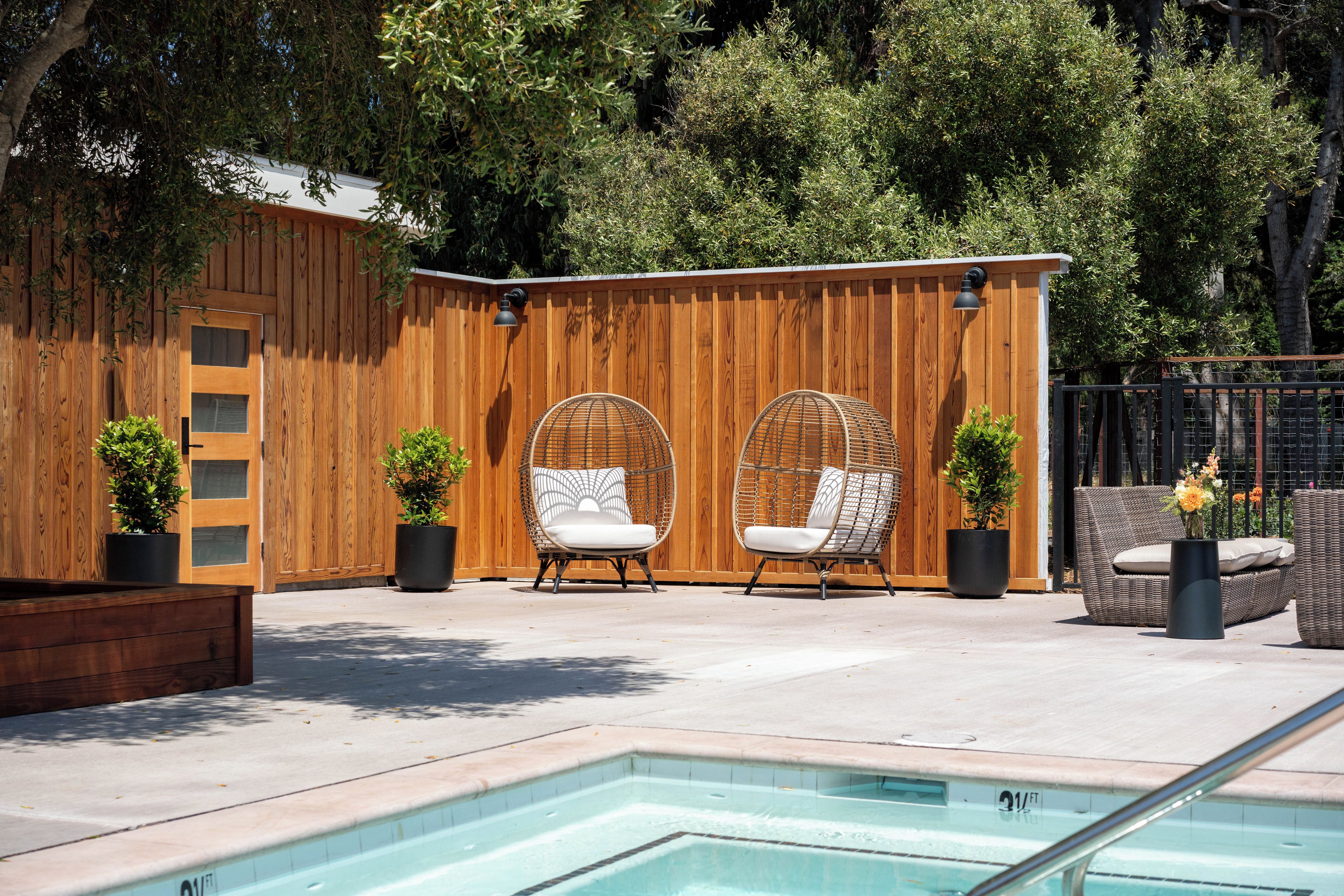 Un área de piscina con dos sillas de mimbre, una terraza de madera y una pequeña estructura de madera con una escalera.