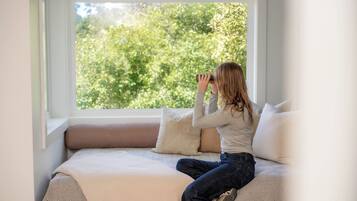 Une personne assise sur un lit, devant une fenêtre donnant sur des arbres.