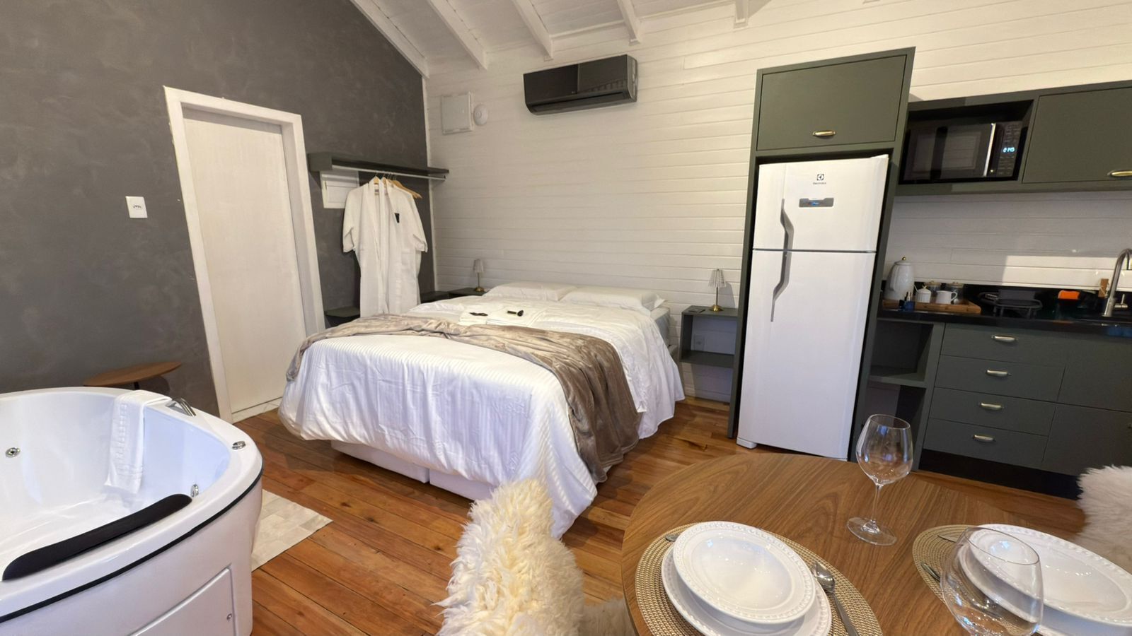 Quarto com cama, banheira branca, mesa de jantar com pratos e uma taça de vinho, e área de cozinha com geladeira e micro-ondas.