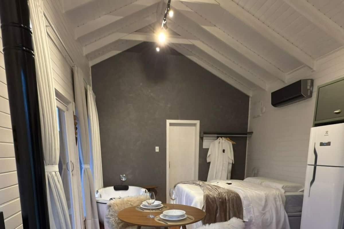 A cozy attic bedroom with a wooden floor, a bed, a small table with chairs, a heater, and a refrigerator.