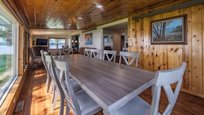 Een kamer met houten lambrisering, een grote eettafel, witte stoelen en een televisie op de achtergrond.