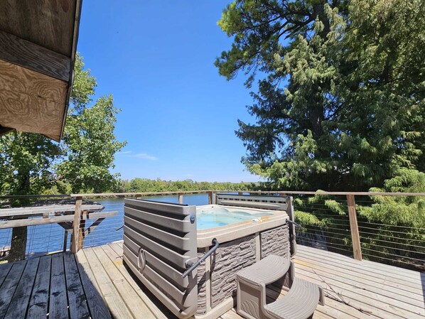 A wooden deck with a hot tub, a stone wall, and a view of a lake surrounded by trees.
