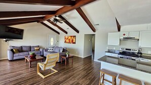 Un'ampia zona giorno con soffitto in legno, un divano grigio, un tavolino da caffè in legno e una cucina con mobili bianchi ed elettrodomestici in acciaio inossidabile.