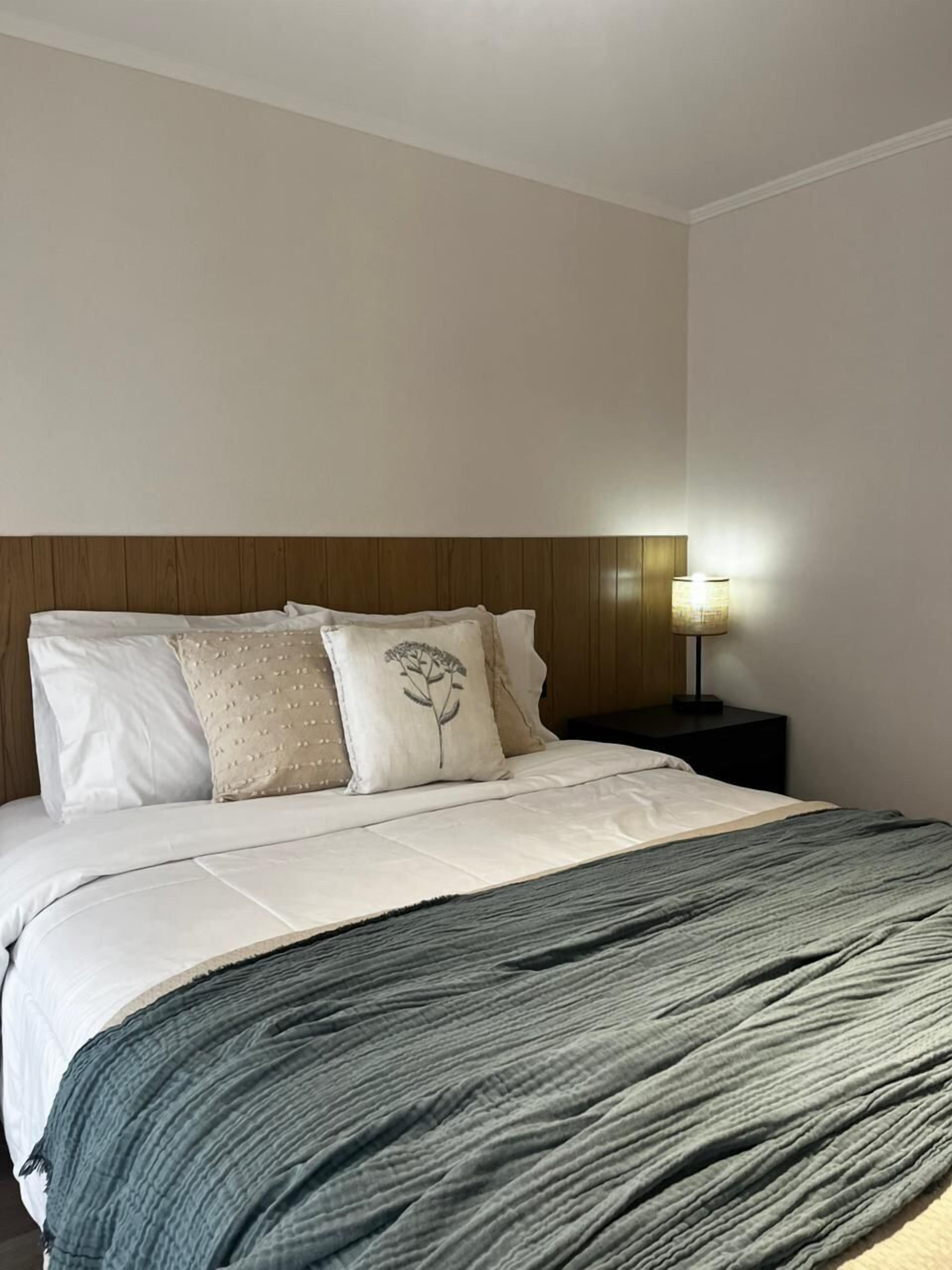 A neatly made bed with a grey blanket, a wooden headboard, and a bedside lamp.