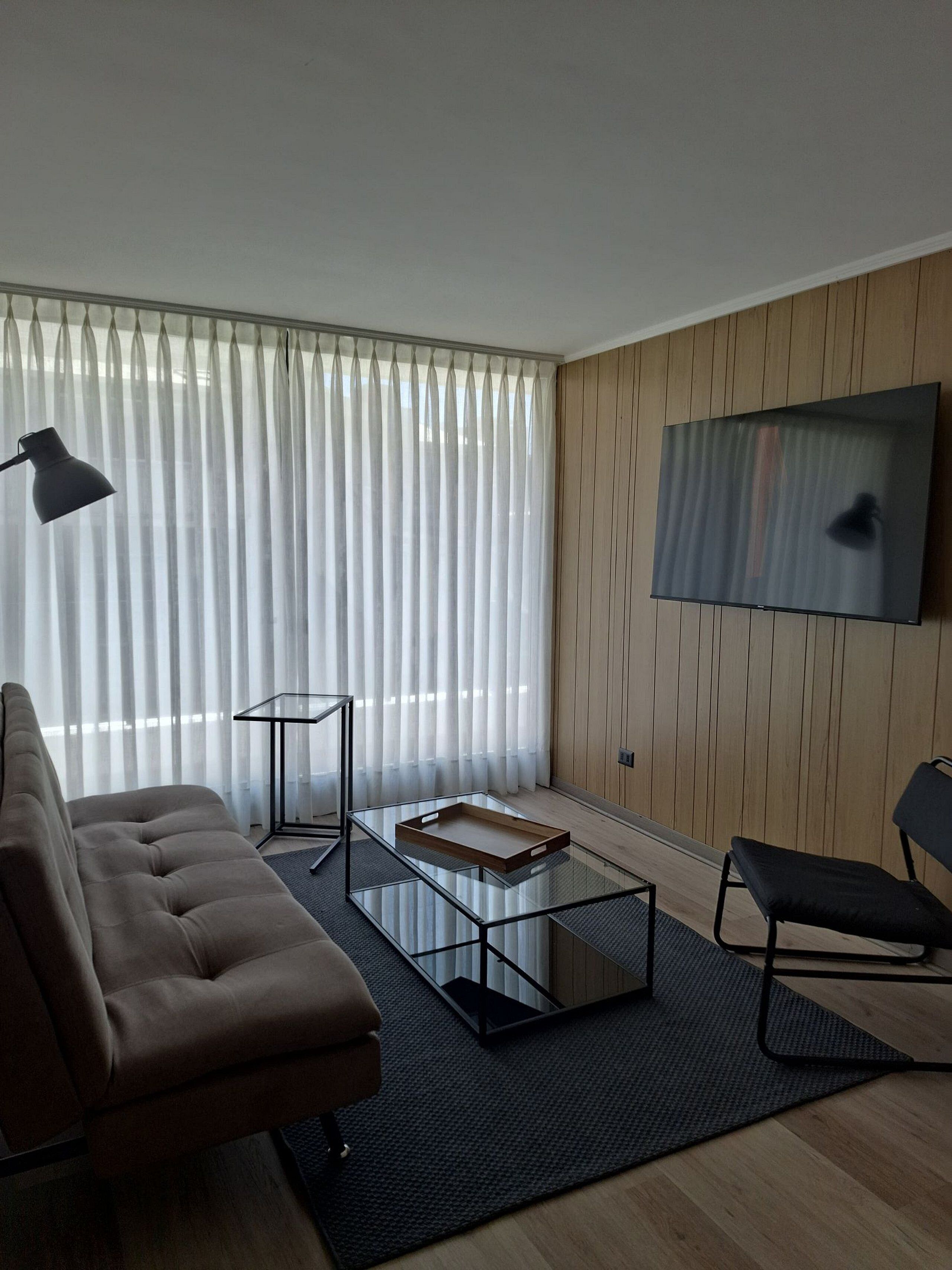 A modern living room with a sofa, a coffee table, a TV mounted on the wall, and sheer curtains.
