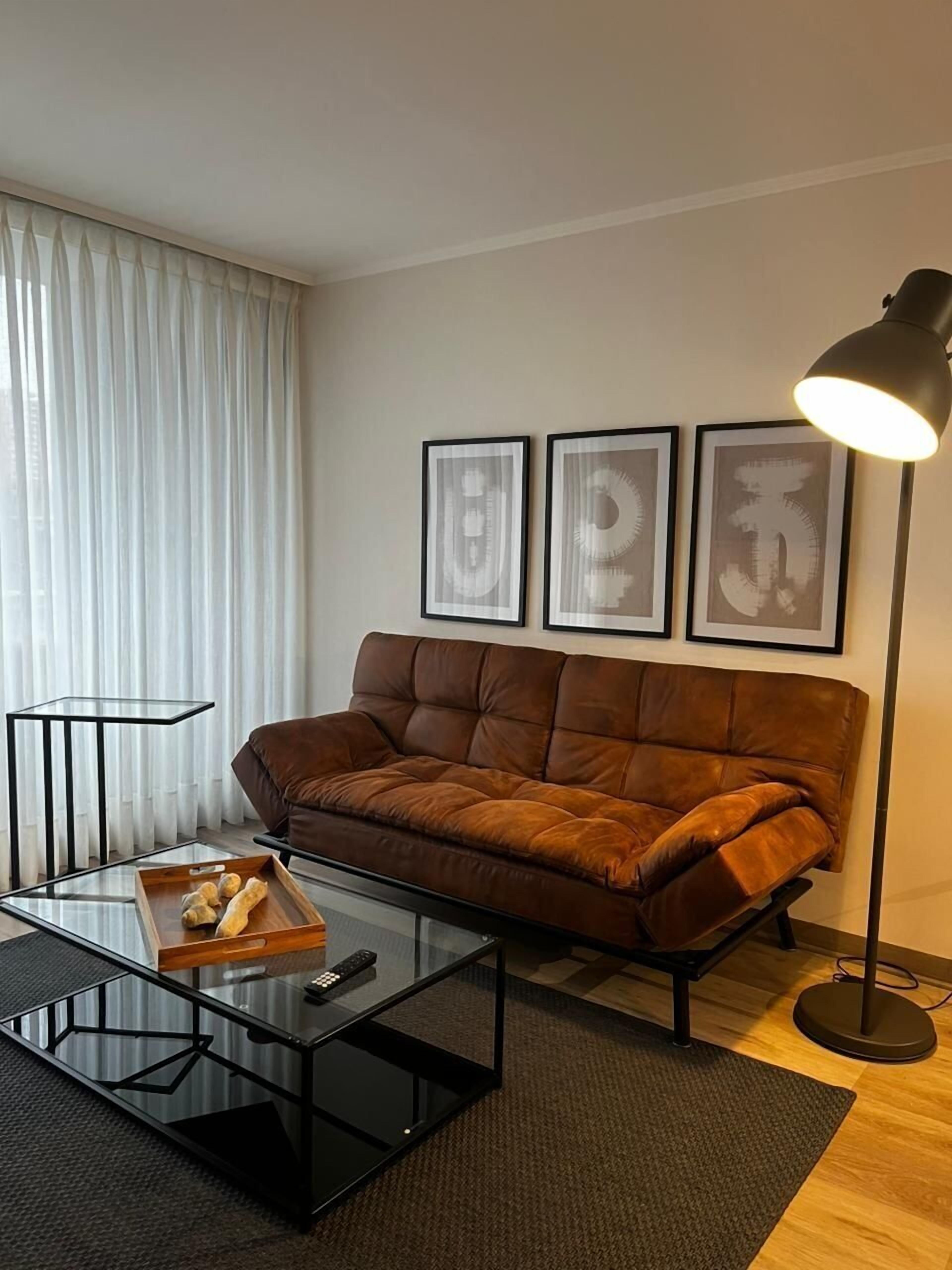 Una sala moderna con un sofá de cuero marrón, una mesa de centro de vidrio con una bandeja de pan, una lámpara de pie negra y tres obras de arte abstracto enmarcadas en la pared.