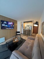 Una sala de estar moderna con un televisor de pantalla plana grande, una mesa de centro de vidrio, un sofá color beige y una zona de cocina al fondo.