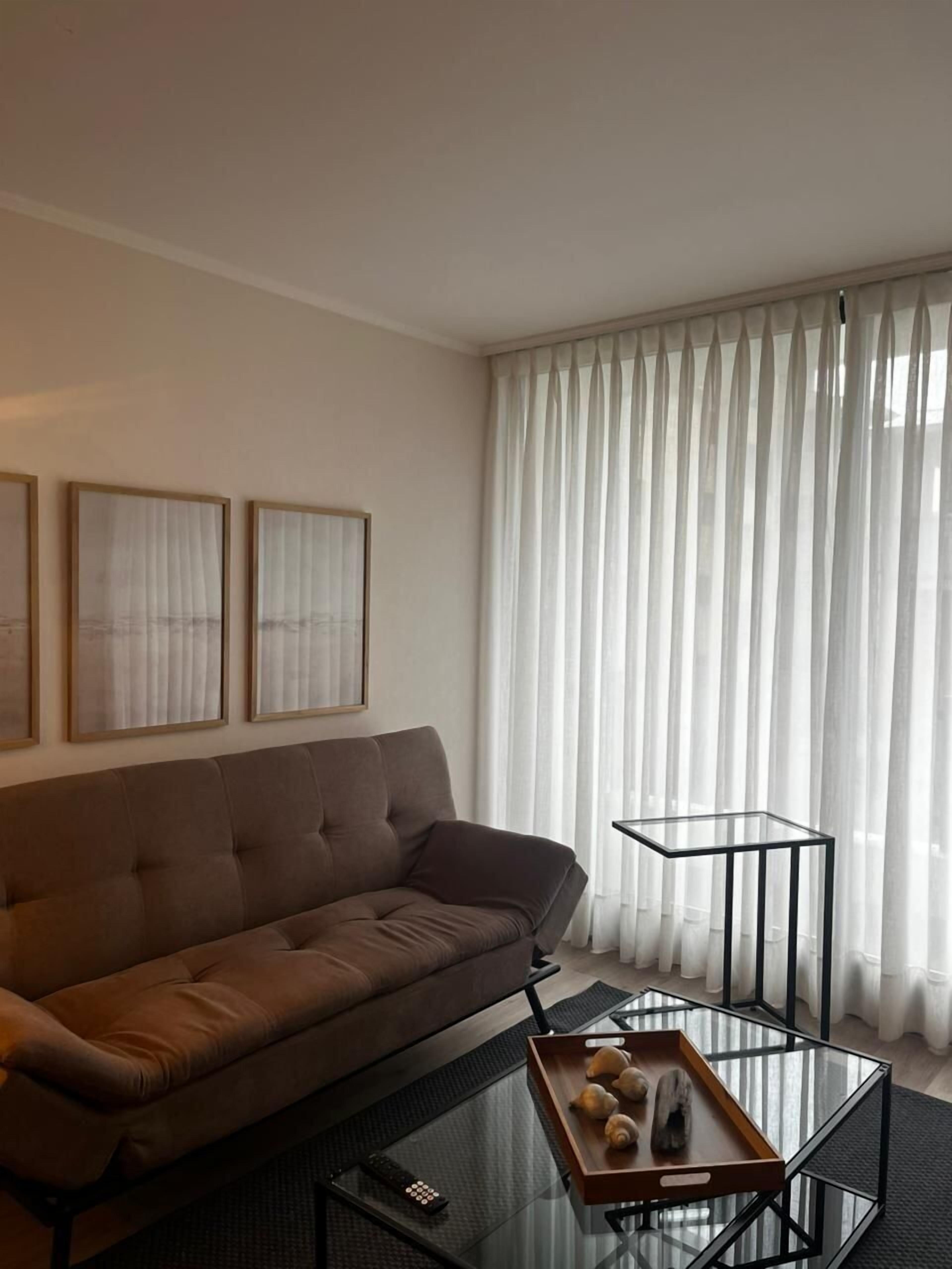 A modern living room with a brown sofa, a glass coffee table with a tray, and sheer curtains.