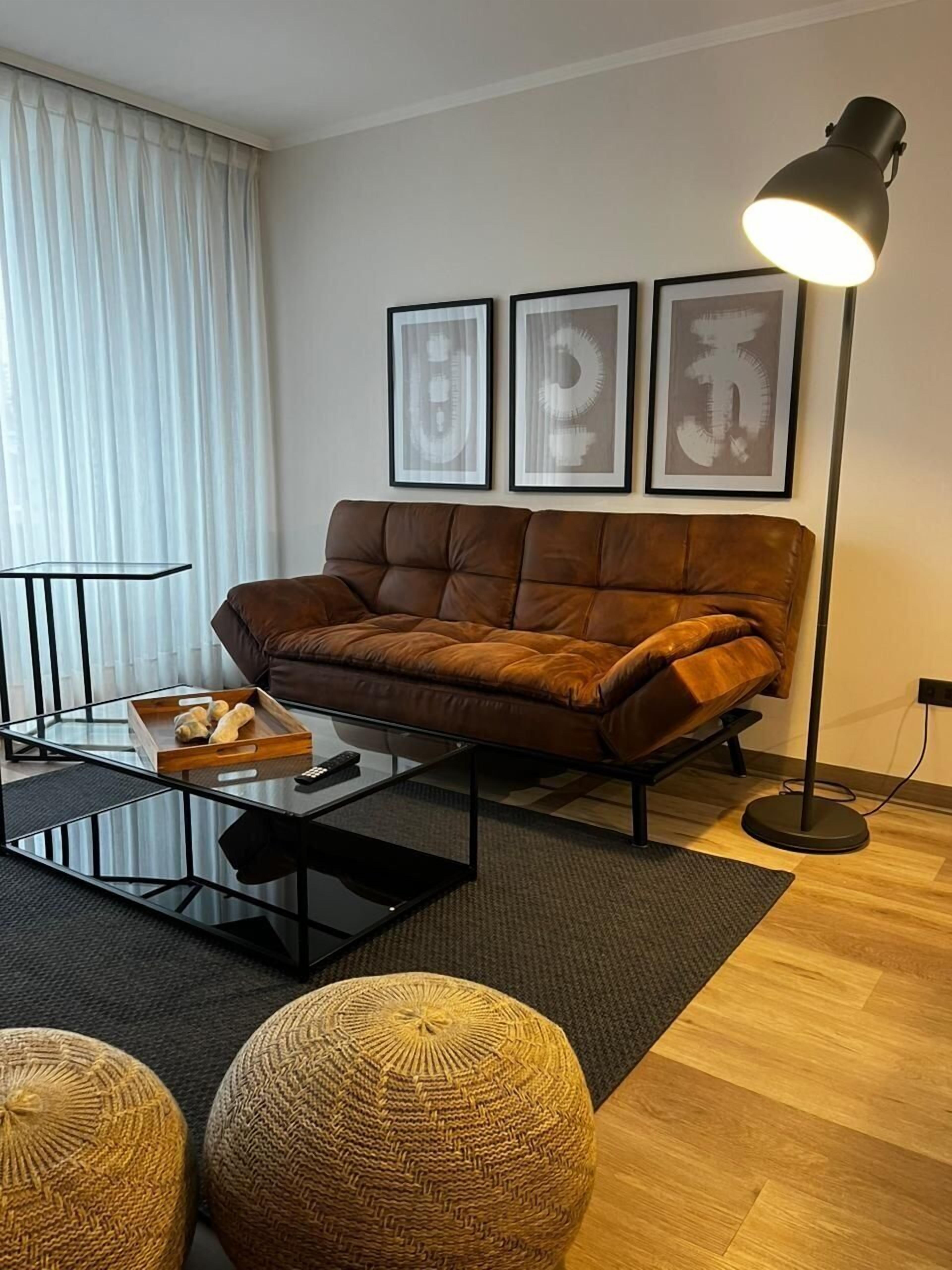 Un moderno salón con un sofá de cuero marrón, una mesa de centro de vidrio y una lámpara de pie. Hay tres obras de arte abstractas enmarcadas en la pared.