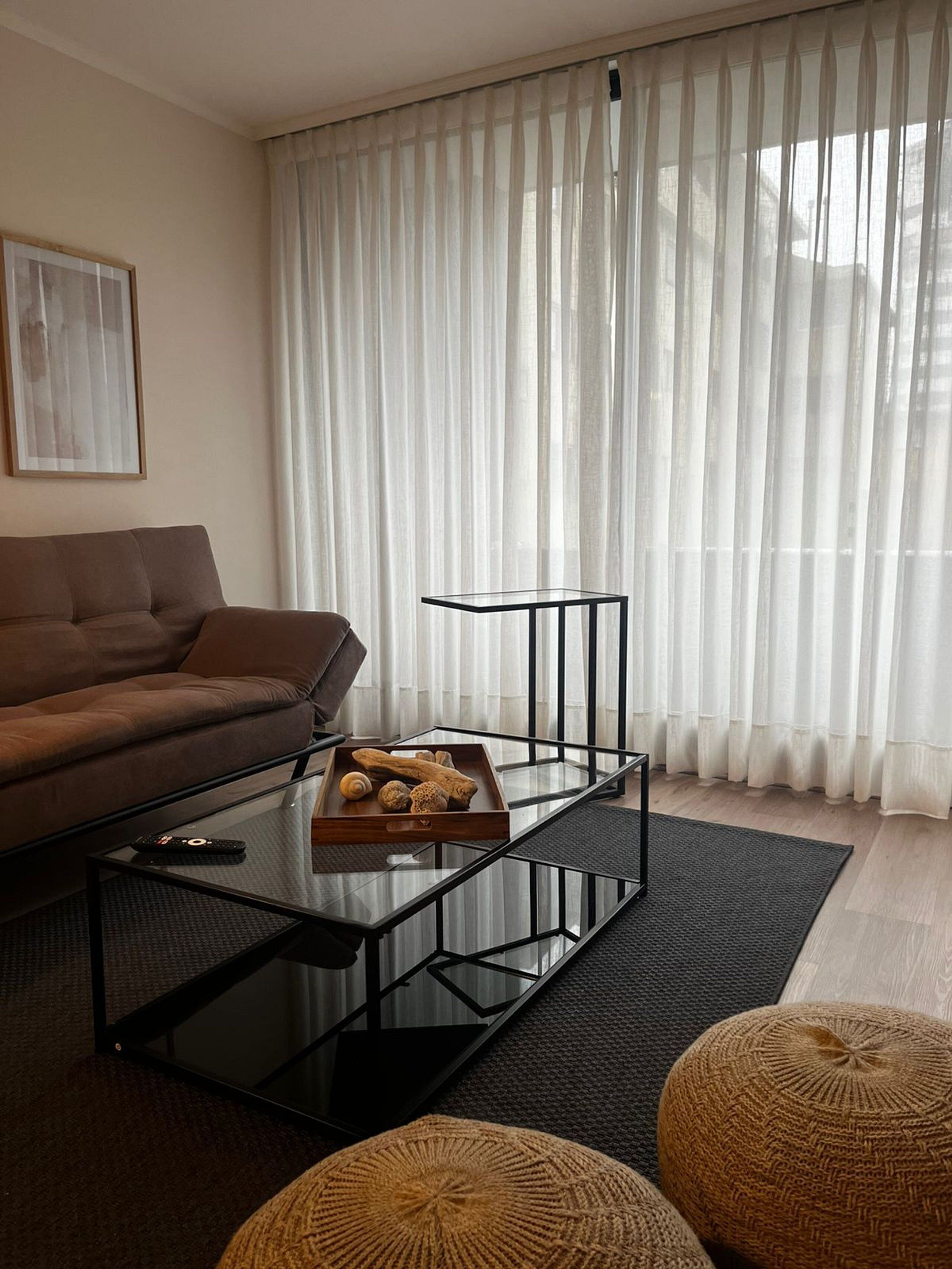 Una sala de estar moderna con un sofá marrón, una mesa de centro de vidrio con una bandeja de bocadillos y un ventanal con cortinas translúcidas.