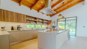 A modern kitchen with wooden cabinets, a central island, and a large window providing a view of greenery. - Casa Nimbu - Hacienda Pinilla (Hacienda Pinilla)