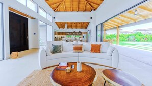 A modern living room with a curved white sofa, a wooden coffee table, and a kitchen in the background. - Casa Nimbu - Hacienda Pinilla (Hacienda Pinilla)