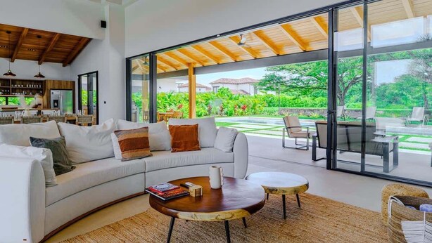 A modern living room with a white sofa, two round coffee tables, and a large glass door leading to an outdoor patio with a view of greenery and a pool. - Casa Nimbu - Hacienda Pinilla (Hacienda Pinilla)