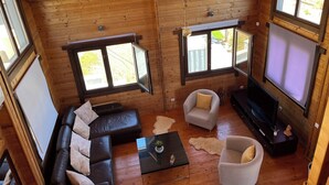 Interior acollidor d'una cabana de fusta amb un sofà de cuir, una poltrona blanca, una taula de centre de vidre i una televisió de pantalla plana.