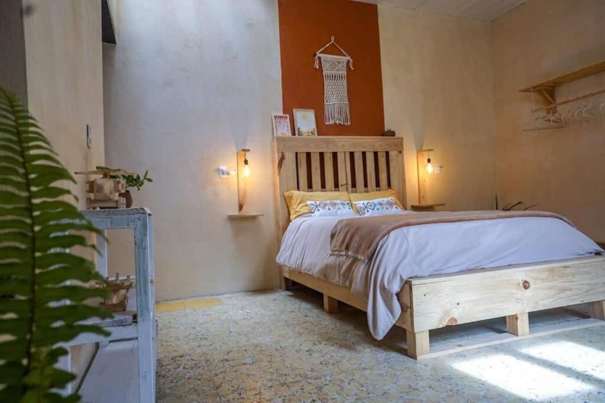 A bedroom with a wooden bed, two bedside lamps, a macrame wall hanging, and a small table with a plant.