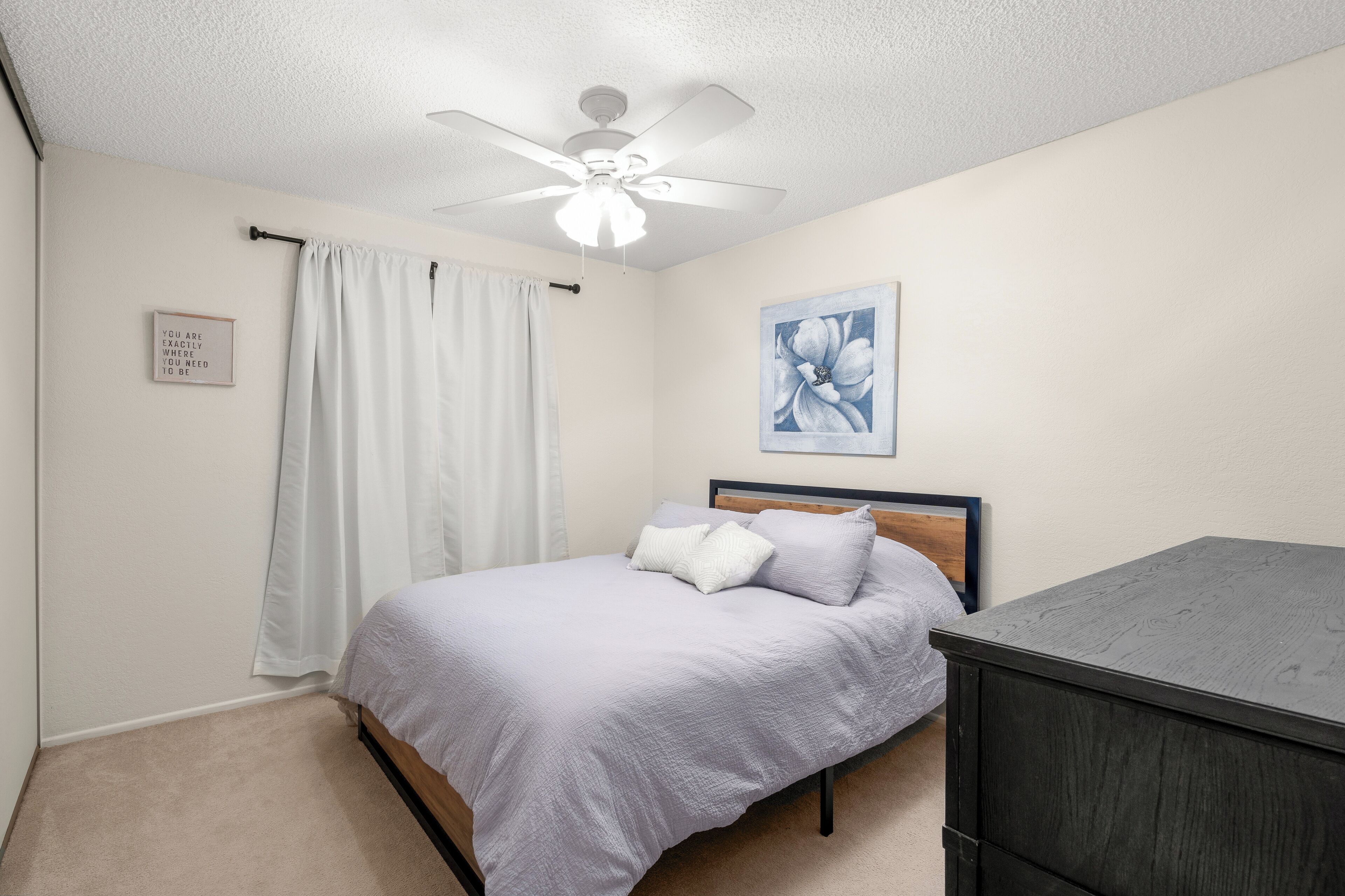 A bedroom with a bed, a ceiling fan, a painting, and a dark wooden dresser.