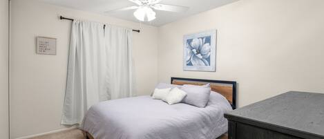 A bedroom with a bed, a ceiling fan, a painting, and a dark wooden dresser.