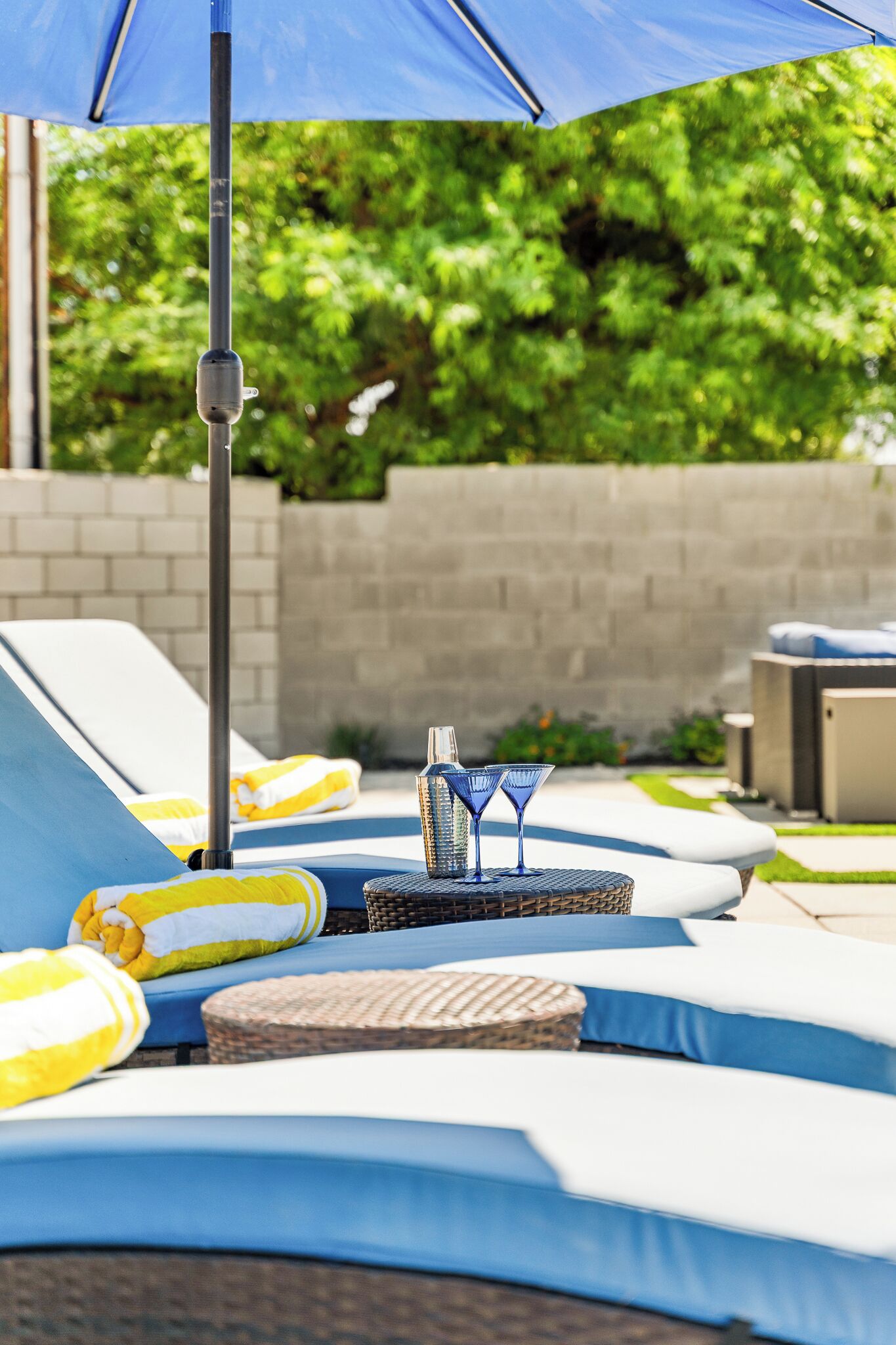 A poolside area with lounge chairs, a table with drinks, and a blue umbrella.