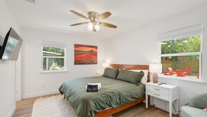 Une chambre à coucher comprenant un lit, une table de chevet, un bureau, un ventilateur de plafond, une fenêtre et un téléviseur.