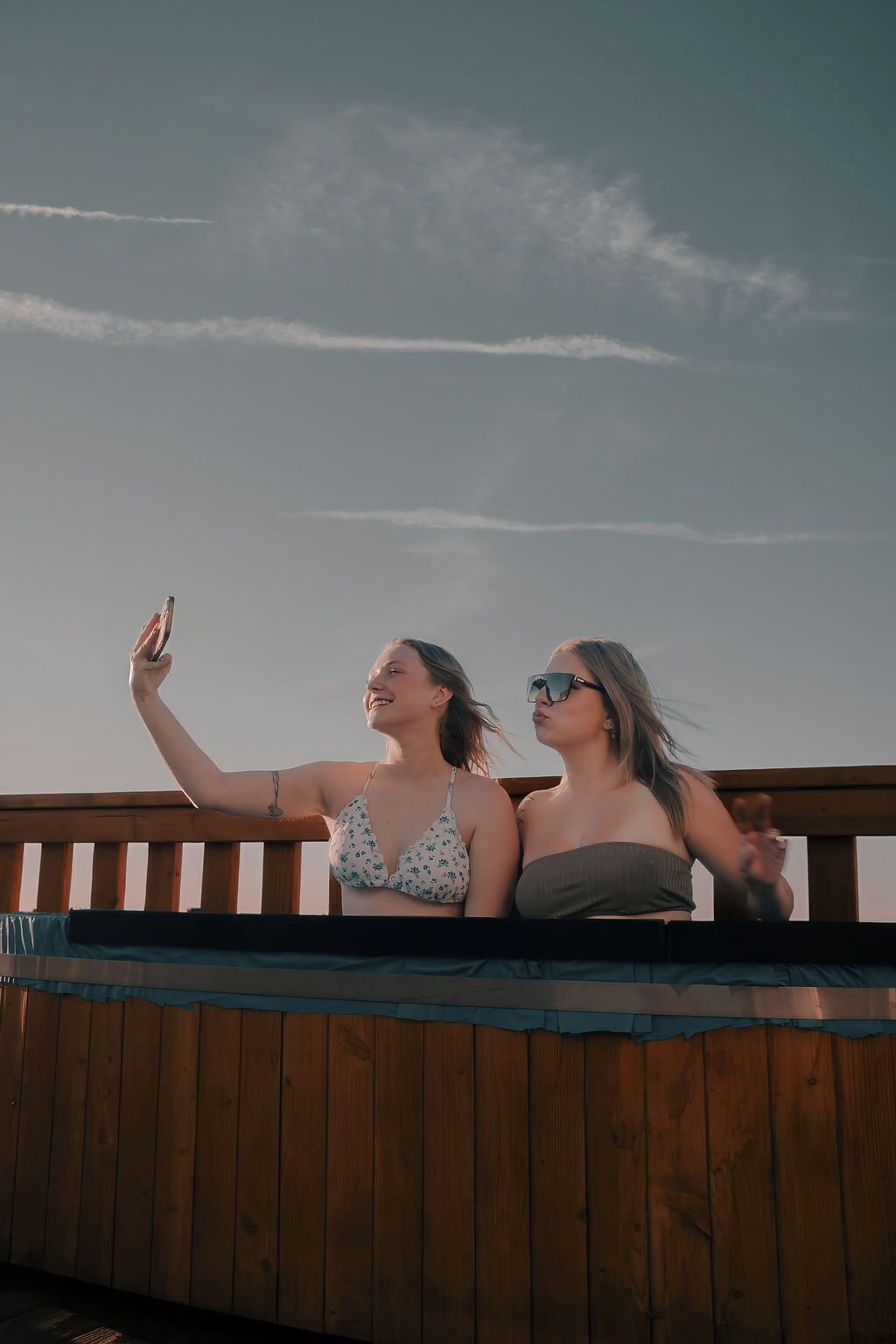 Zwei Personen machen ein Selfie auf einer Holzterrasse, im Hintergrund ist ein klarer Himmel zu sehen.