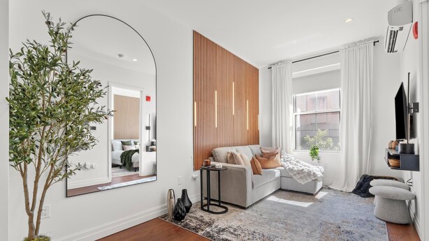 Una sala de estar moderna con un espejo grande, una pared de acento de madera, un sofá gris y un ventanal con cortinas blancas.