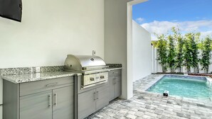 Een moderne buitenkeuken met een ingebouwde barbecue, een terras met stenen tegels en een zwembadgedeelte met bamboeplanten.