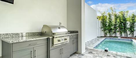 A modern outdoor kitchen with a built-in grill, a stone-tiled patio, and a pool area with bamboo plants.