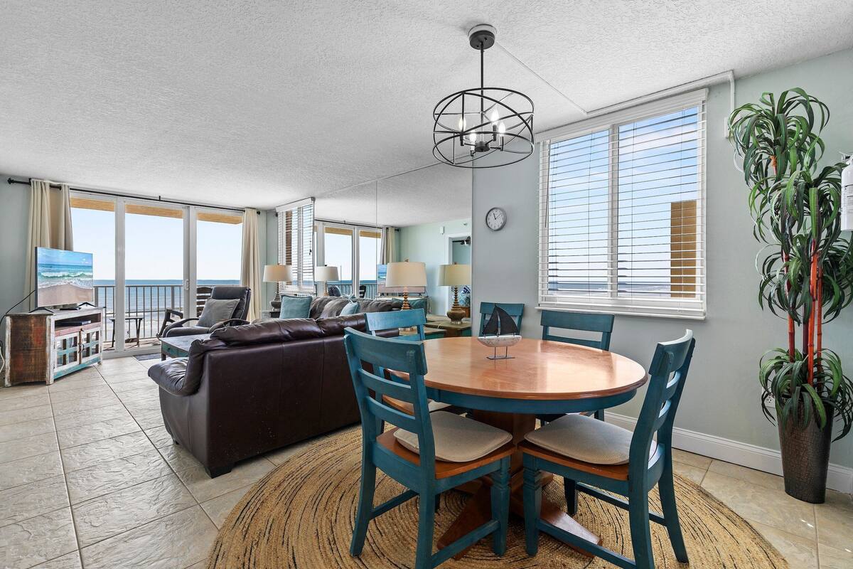 A spacious living room with a large window, a dining area with a wooden table and chairs, a brown leather sofa, and a potted plant.