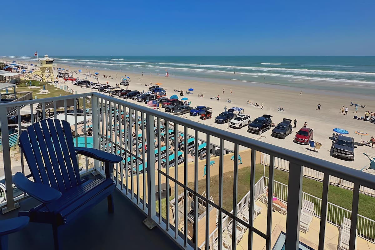 A balcony with a view of a beach, a pool, and several lounge chairs.