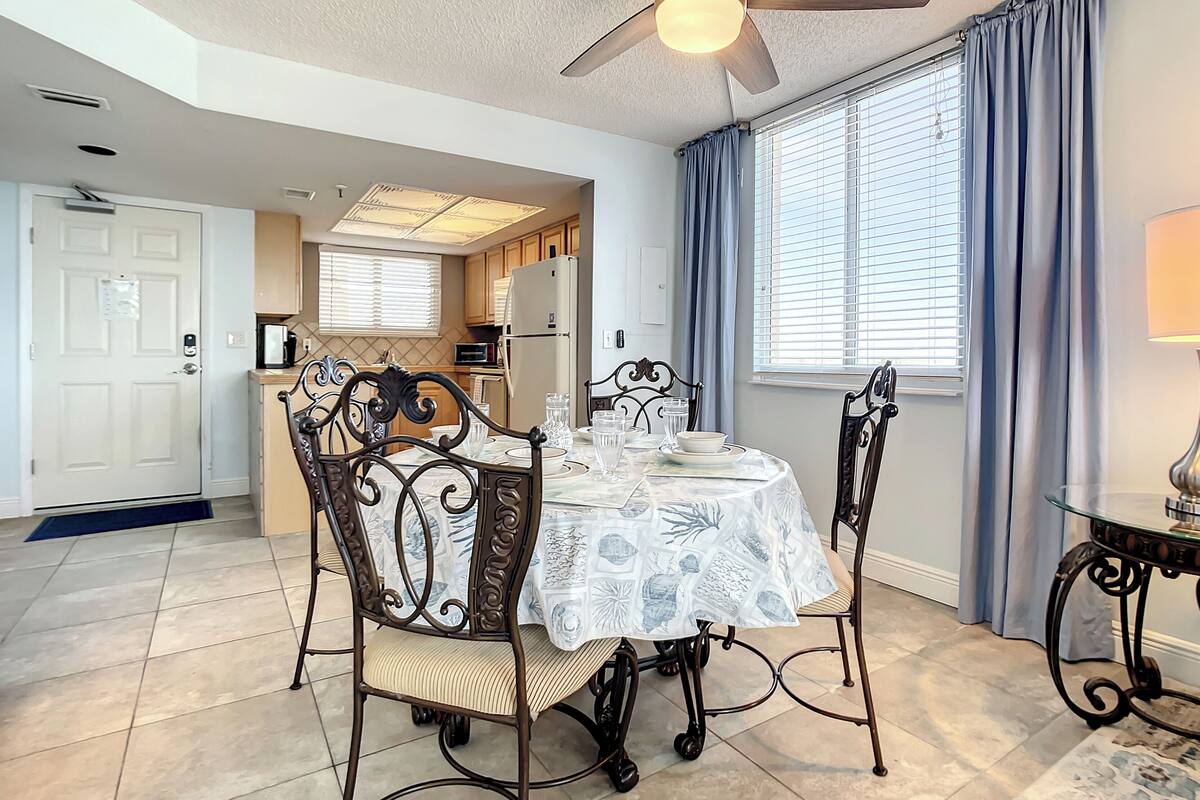 A dining area with a table set for four, surrounded by ornate chairs, a ceiling fan, and a view of the kitchen through a window with curtains.
