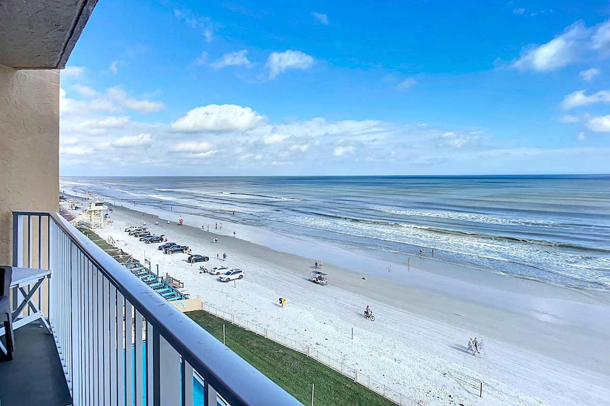 A balcony overlooking a beach with a clear sky, a sandy shore, and a few people and vehicles.