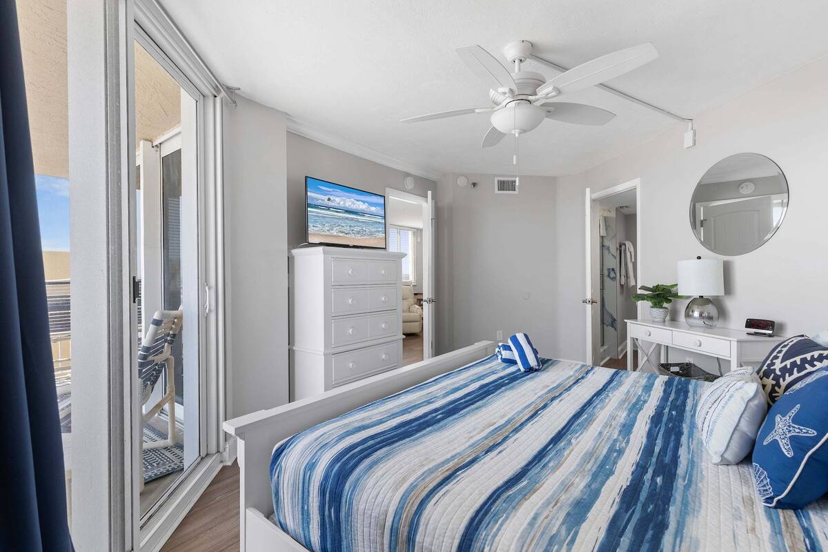 Un dormitorio con una cama, ventilador de techo, televisor, cómoda y puerta que da a otra habitación.