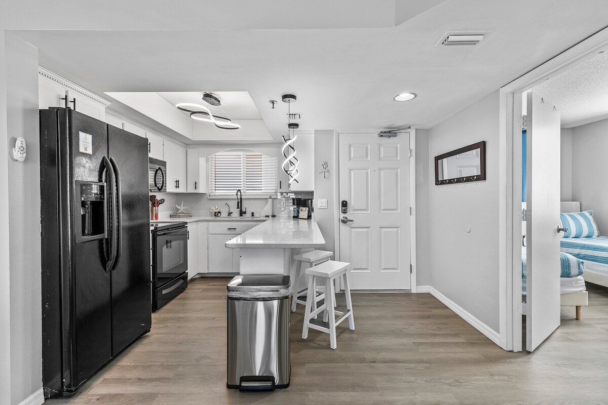 A modern kitchen with a black refrigerator, white cabinets, a stainless steel trash can, and a bar stool.