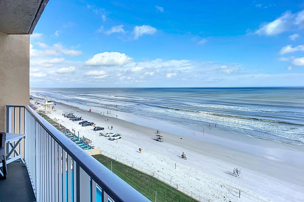A balcony overlooking a beach with a clear sky, a sandy shore, and a few people and vehicles.