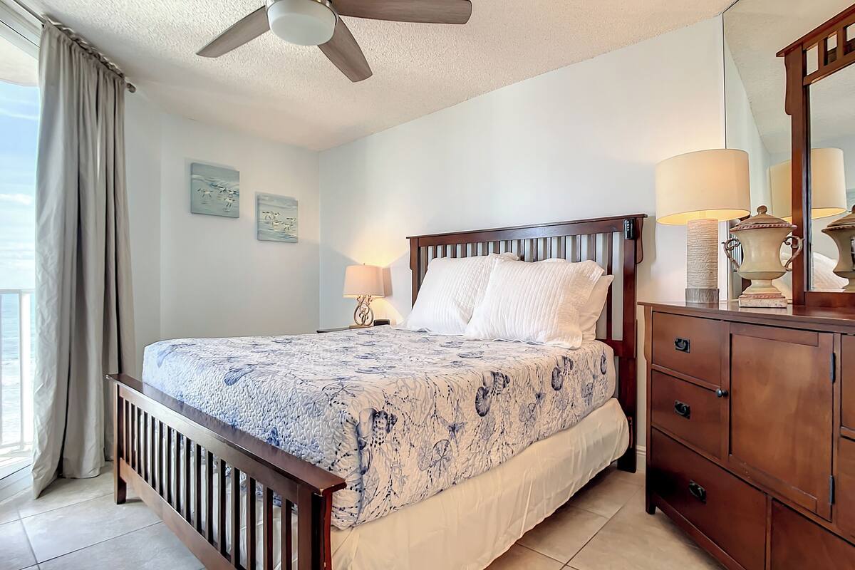 Un dormitorio con cama, cómoda de madera, ventilador de techo y ventana con cortinas.