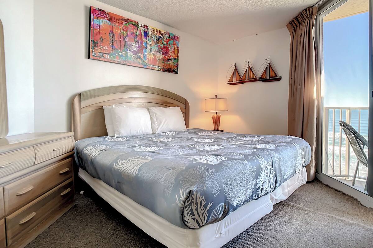 Habitación de hotel con cama, mesita de noche, lámpara, un cuadro y vista al mar.
