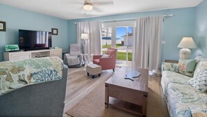 A living room with a television, two sofas, a coffee table, and a chair. There is a view of a balcony with a railing. - Oceanside Village C1 (New Smyrna Beach)