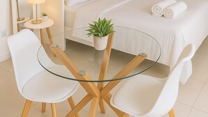 Una habitación de hotel moderna con una mesa de vidrio, dos sillas blancas, una cama con ropa de cama blanca, una pequeña planta en maceta y una lámpara de noche.