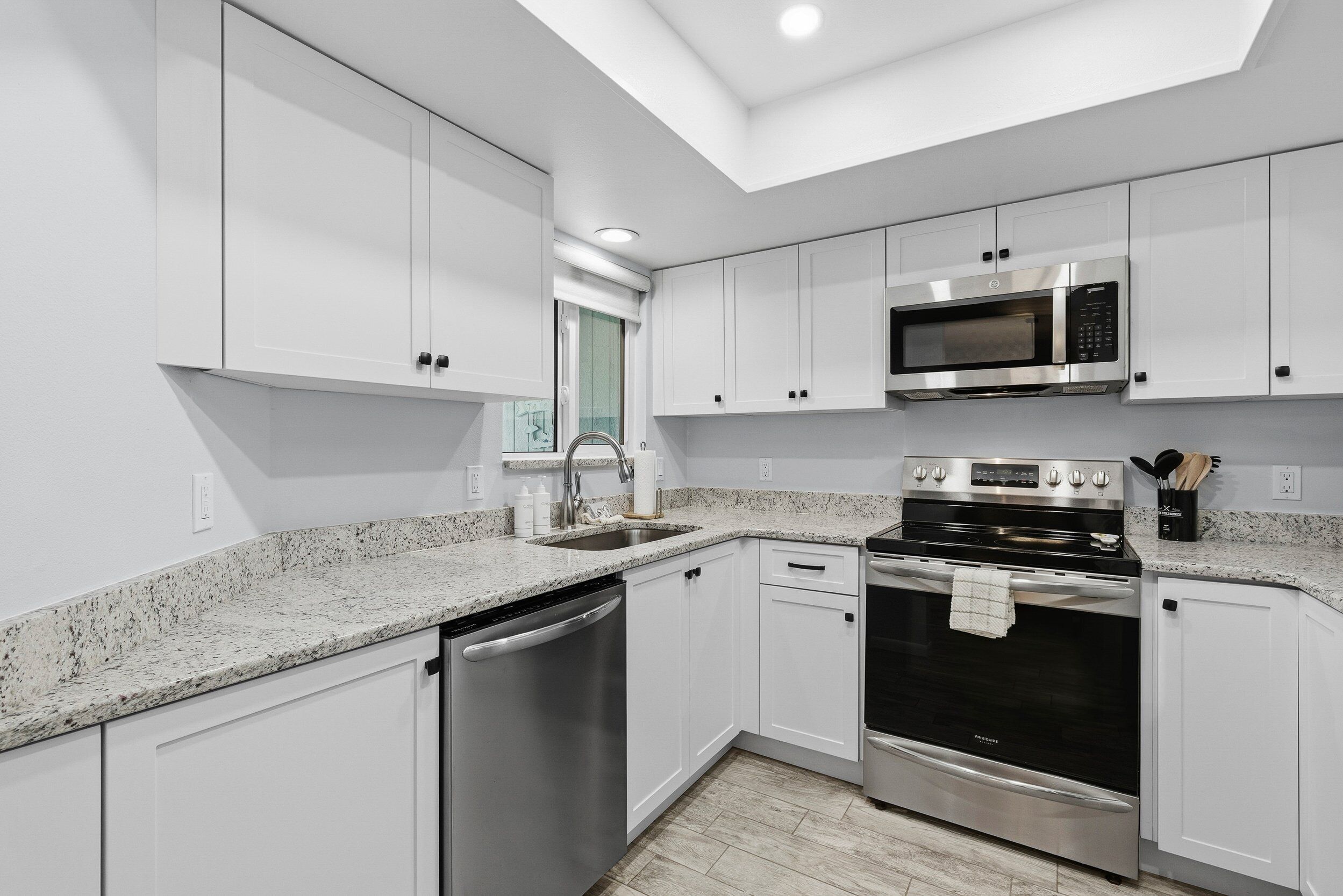 A modern kitchen with white cabinets, stainless steel appliances, a granite countertop, and a skylight.