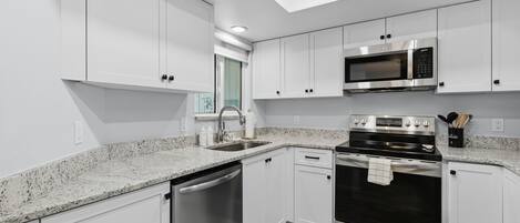 A modern kitchen with white cabinets, stainless steel appliances, a granite countertop, and a skylight.