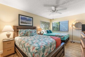 Une chambre avec deux lits, un ventilateur de plafond, une table de chevet avec une lampe, une commode et un tableau encadré au mur.