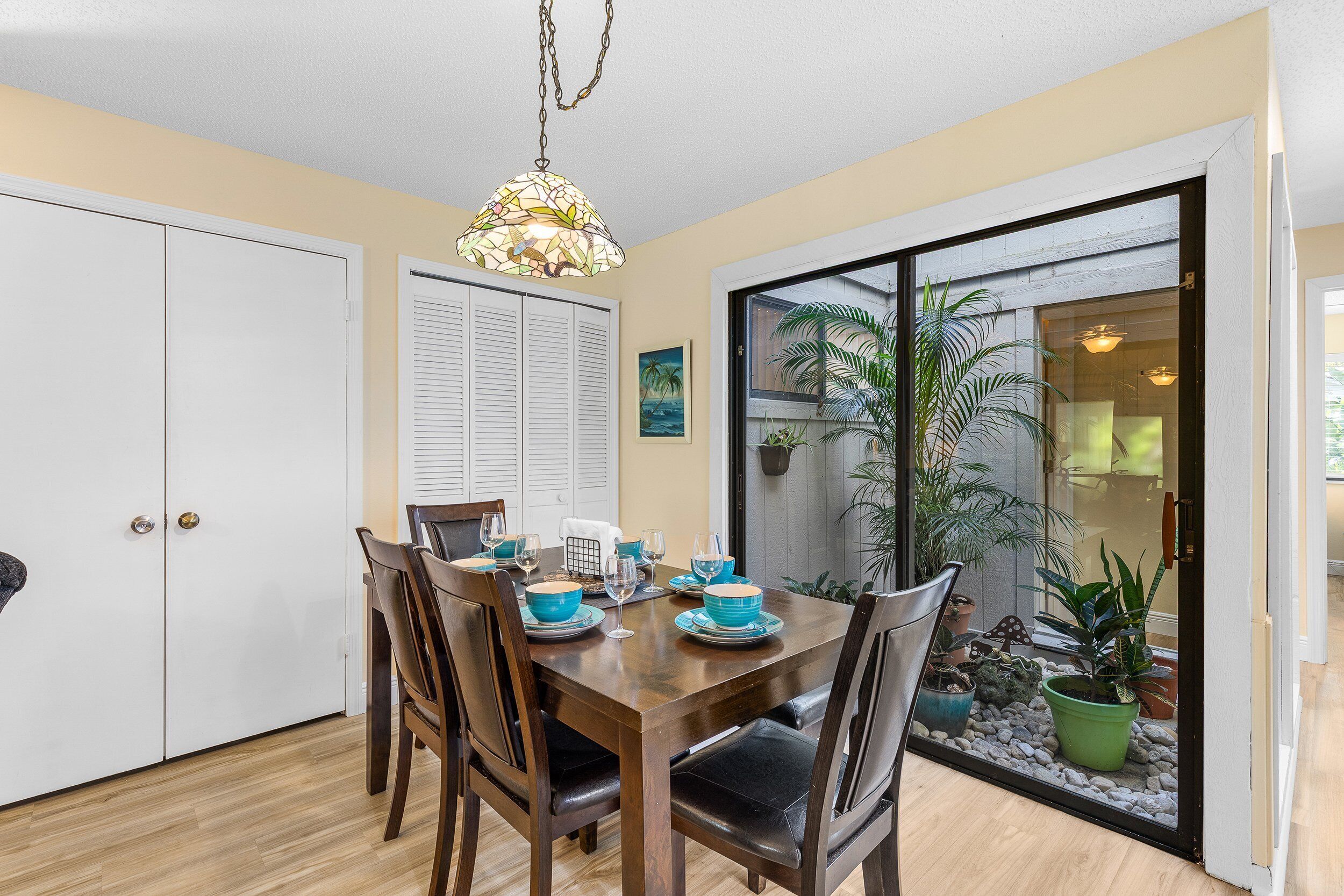 A dining area with a wooden table set for four, surrounded by chairs. There is a white door and a window with blinds on the wall.