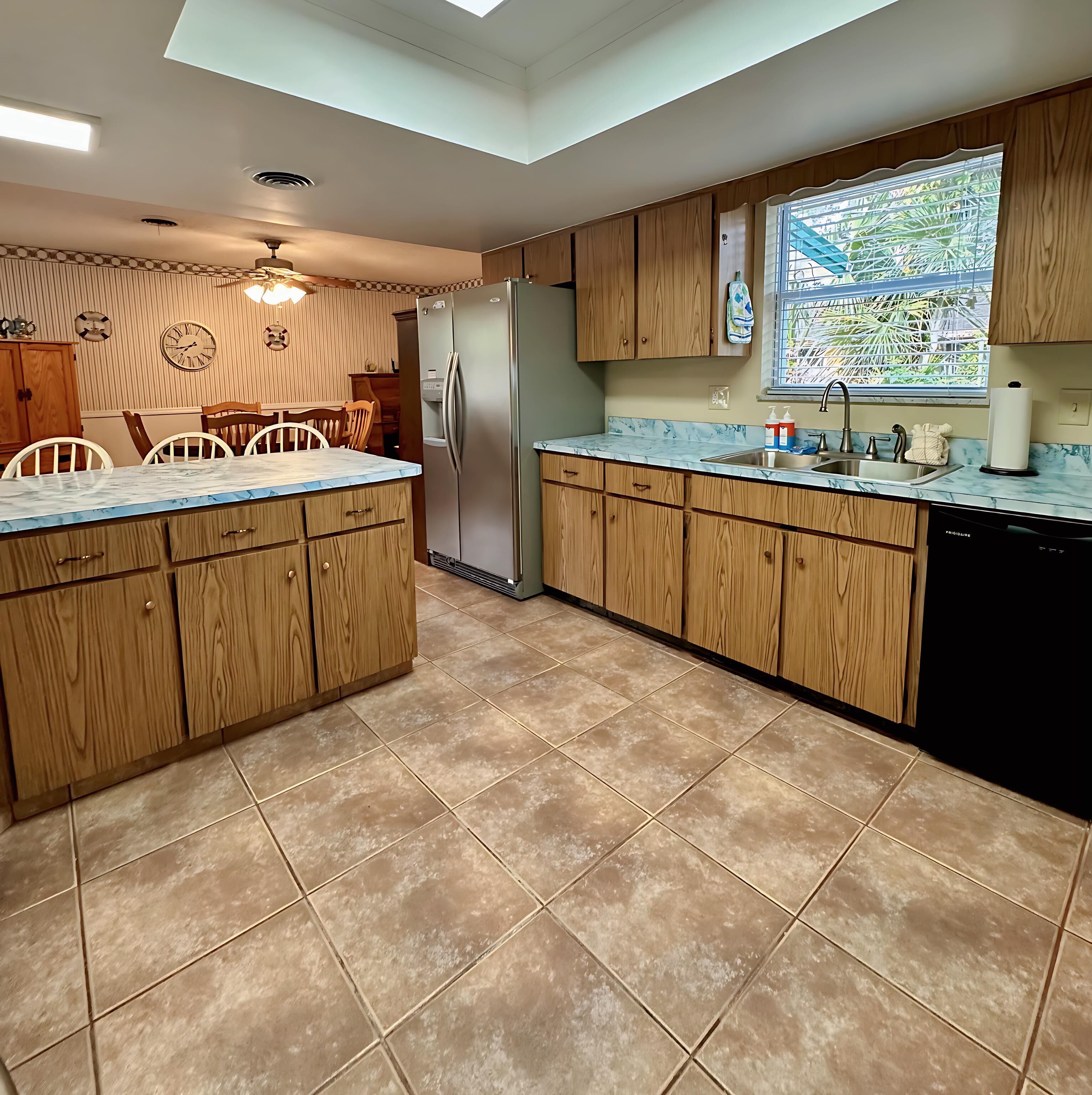 A kitchen with wooden cabinets, a marble countertop, a refrigerator, and a window with a view of greenery.