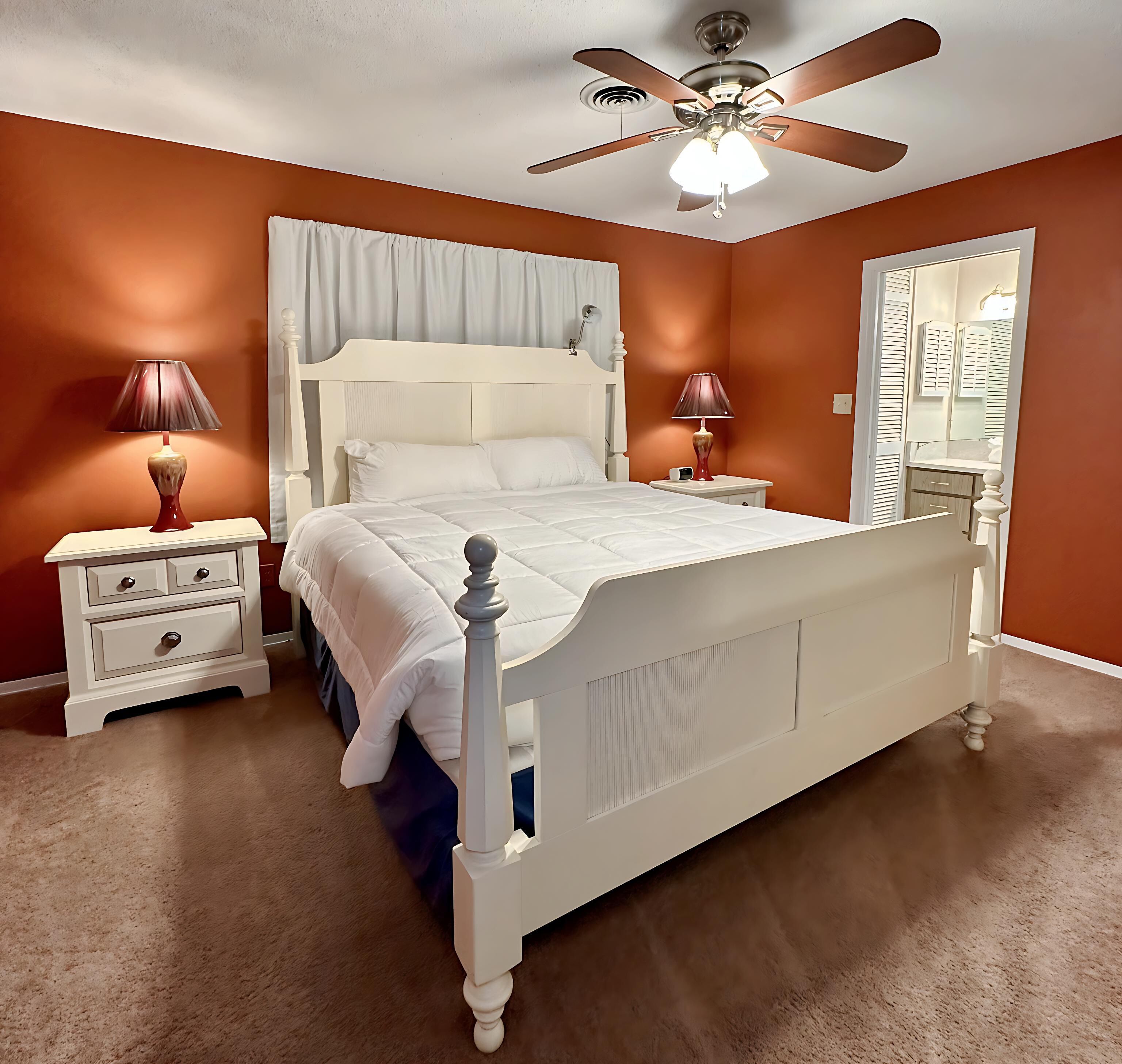 A bedroom with a white bed, two bedside tables with lamps, a ceiling fan, and a large mirror.