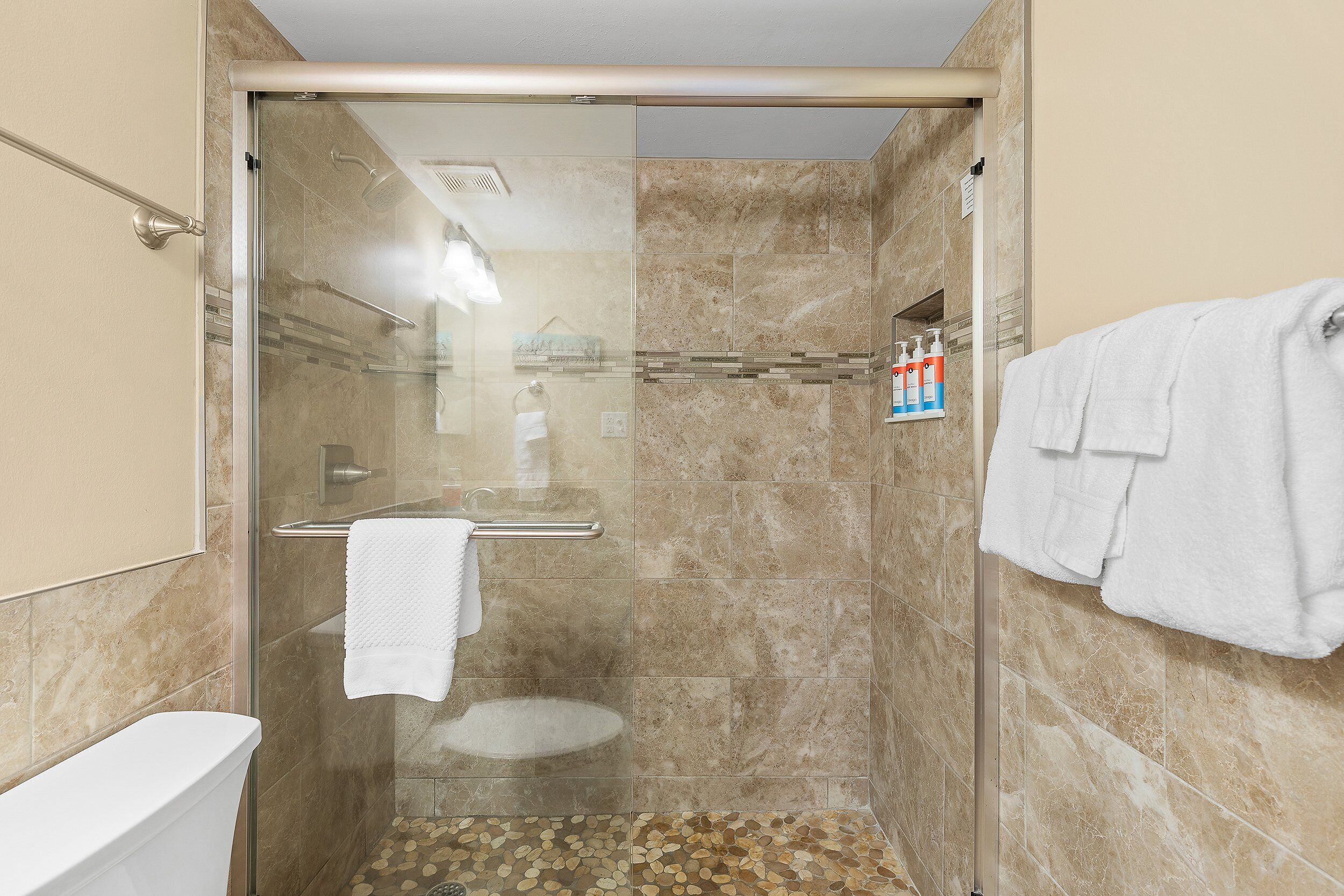 A glass-enclosed shower with beige tiles, a white towel, and toiletries on a shelf.