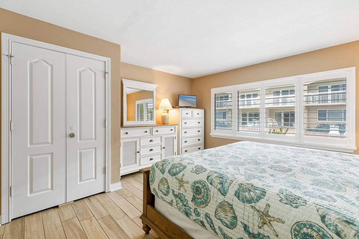 Une chambre à coucher comprenant un lit, une commode, un miroir et une fenêtre donnant sur des immeubles.