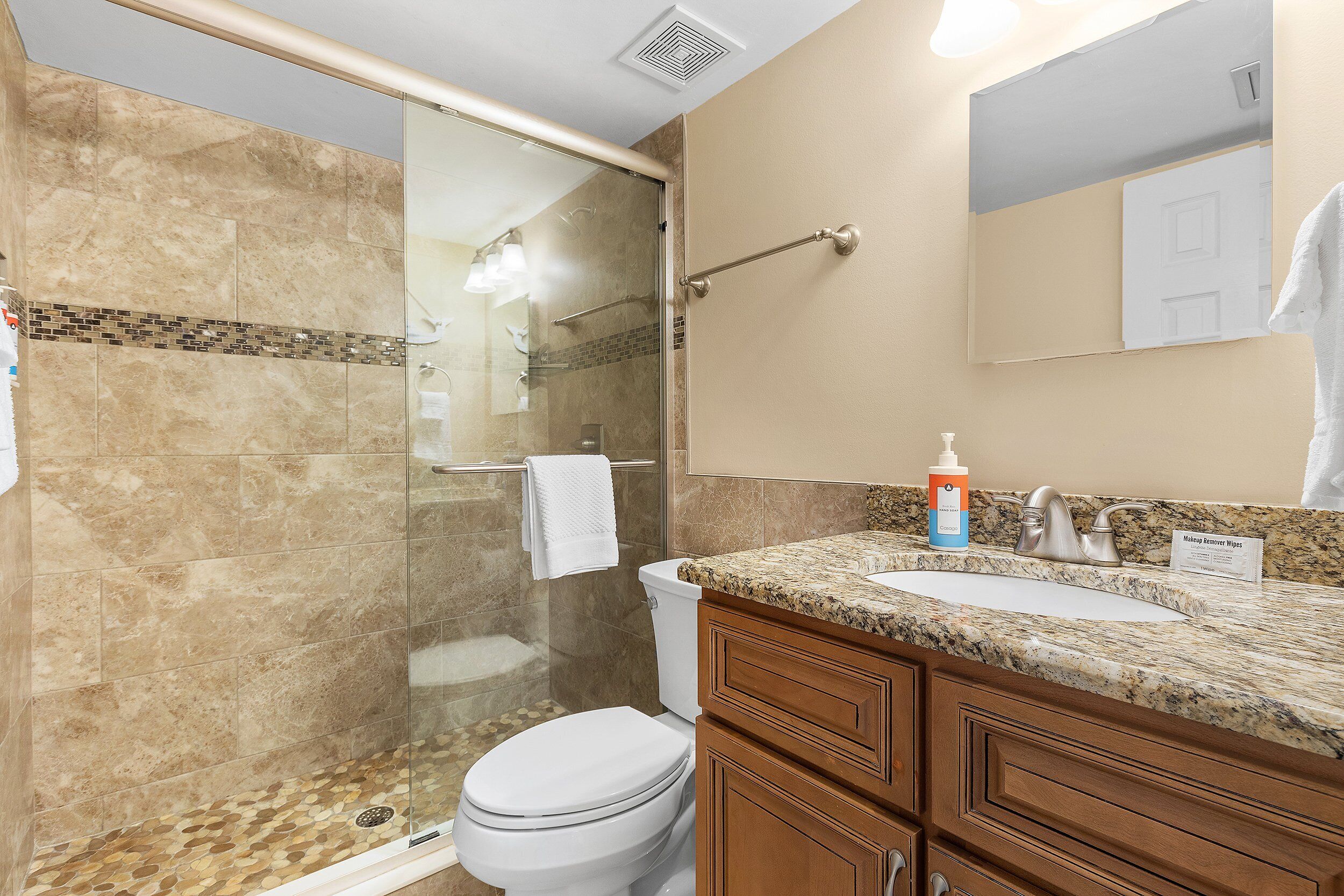 A bathroom with a glass shower enclosure, a toilet, a sink with a granite countertop, and wooden cabinets.