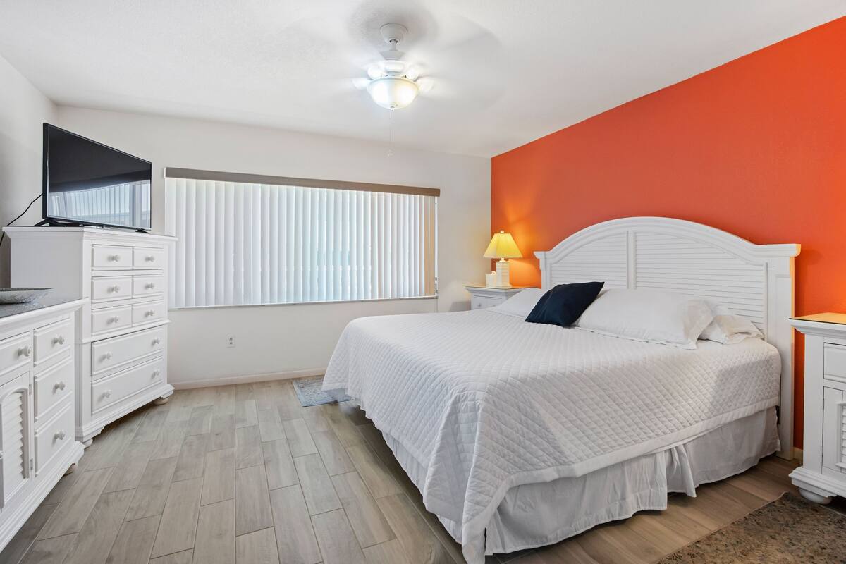 Une chambre à coucher avec un lit, une commode, un téléviseur et une fenêtre avec des stores.