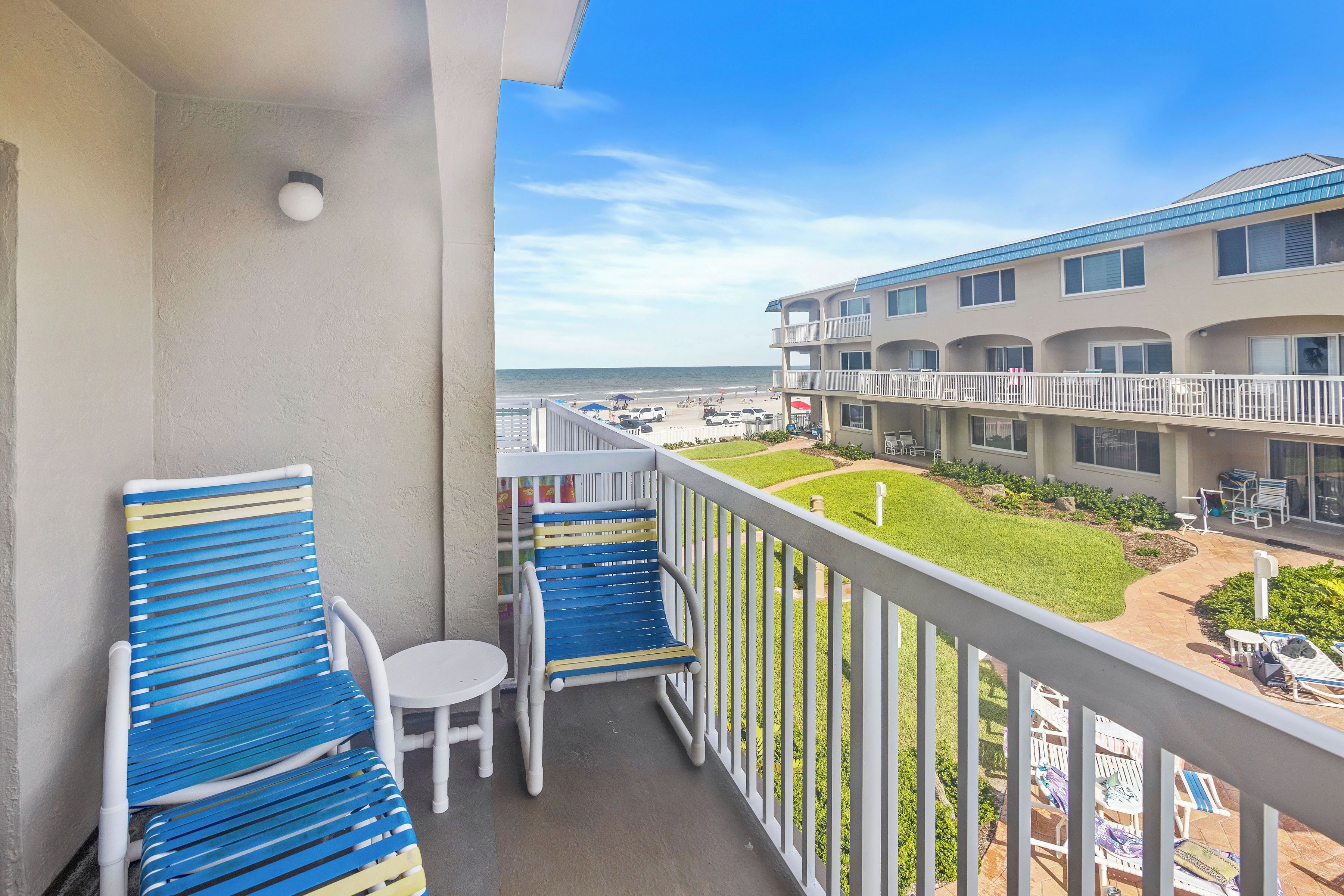 A balcony with two blue chairs and a small table, overlooking a beach and a hotel building with a garden.