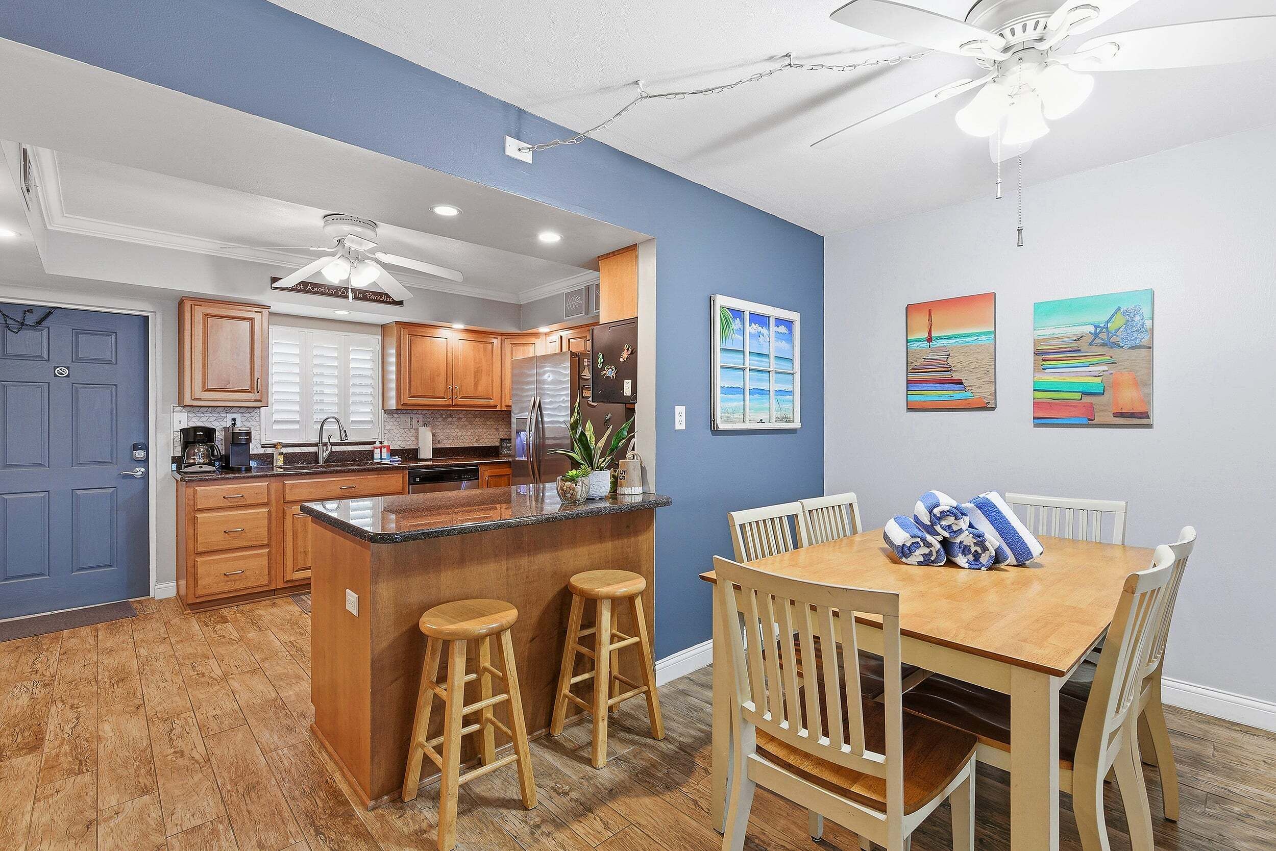 A modern kitchen with wooden cabinets, a central island with stools, a dining area with a wooden table and chairs, and artwork on the walls.